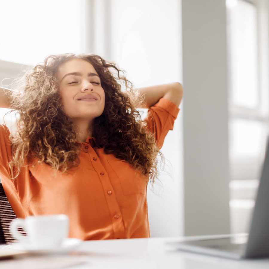 Curly-haired businesswoman leaning back in her chair with eyes closed, taking a relaxing break from work in a sunny, modern office space, free space
